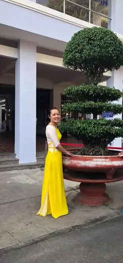 Một cô giáo mặc áo dài vàng đứng trước sảnh một toà nhà.