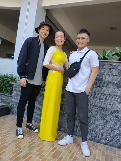 Một cô giáo mặc áo dài vàng đứng giữa hai thanh niên.