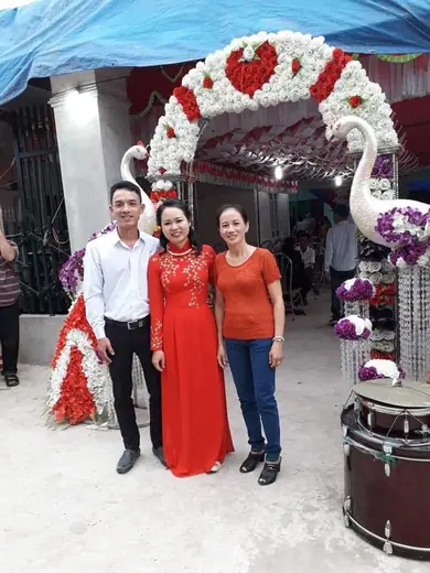 Một phụ nữ mặc áo dài đỏ đứng cùng người thân trước cổng hoa đám cưới.