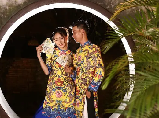 Một đôi nam nữ mặc áo dài gấm vàng hoạ tiết rồng cầm quạt trong cổng tròn.