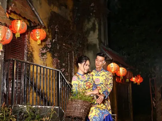 Một đôi nam nữ mặc áo dài gấm vàng ngồi bên bậc thềm treo đèn lồng.