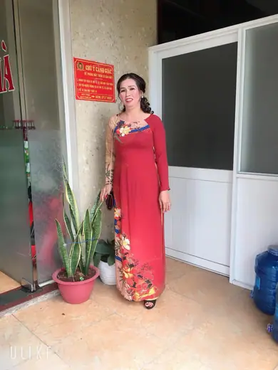 Một phụ nữ mặc áo dài đỏ đô in hoa đứng trước cửa nhà.