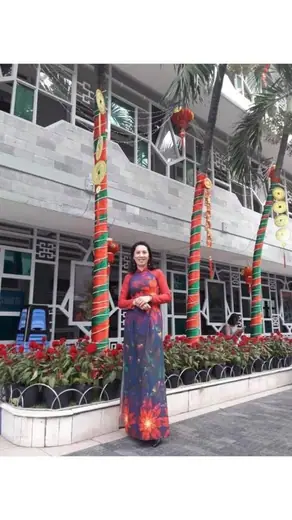 Một phụ nữ mặc áo dài tối màu in hoa đỏ đứng trước toà nhà trang trí Tết.