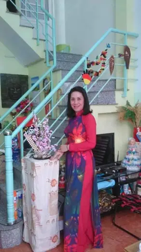 Một phụ nữ mặc áo dài đỏ phối xanh đứng bên cầu thang có cành hoa đào.