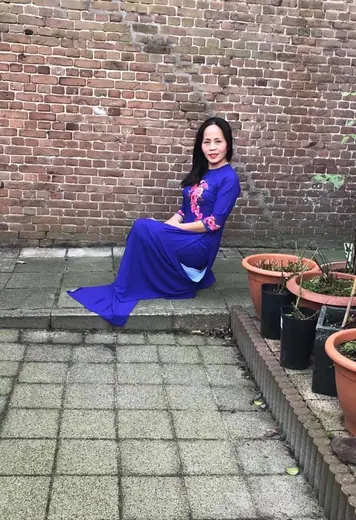 Một phụ nữ mặc áo dài xanh dương thêu hoa ngồi trước bức tường gạch cũ.
