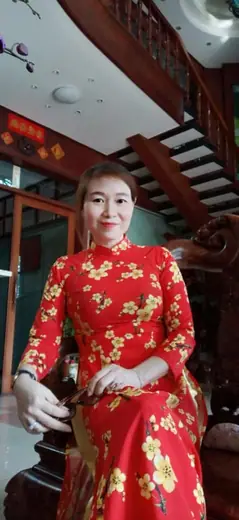 Một phụ nữ mặc áo dài đỏ in hoa mai vàng ngồi trên ghế gỗ.