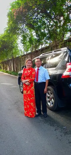 Một phụ nữ mặc áo dài đỏ in hoa mai đứng cùng một người đàn ông bên chiếc ô tô.