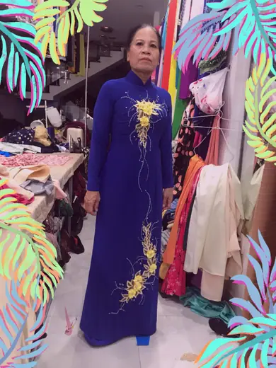 Một phụ nữ lớn tuổi mặc áo dài xanh dương thêu hoa vàng đứng trong tiệm, ảnh có khung trang trí.