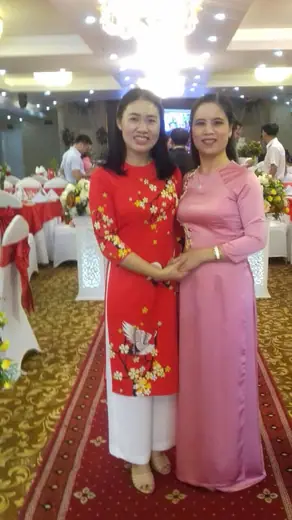 Hai phụ nữ mặc áo dài đỏ in hoa và hồng đứng trong sảnh tiệc cưới.