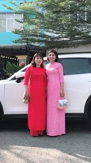 Hai phụ nữ mặc áo dài đỏ và hồng đứng bên chiếc ô tô trắng.