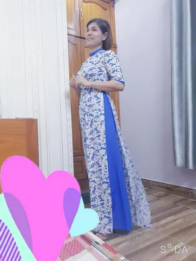 Một phụ nữ mặc áo dài trắng hoa xanh phối tà xanh dương đứng trong nhà, ảnh có hình dán trang trí.