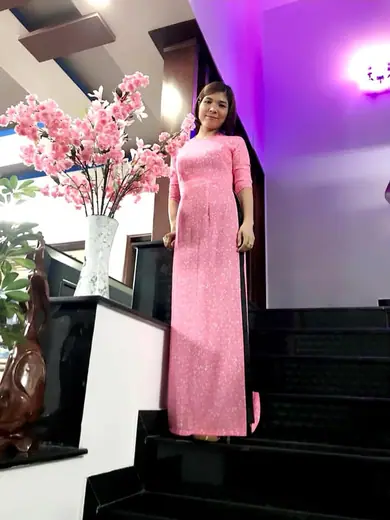 Một phụ nữ mặc áo dài hồng đứng trên cầu thang bên bình hoa đào.