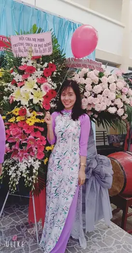 Một cô giáo mặc áo dài trắng hoa tím đứng bên các lẵng hoa.