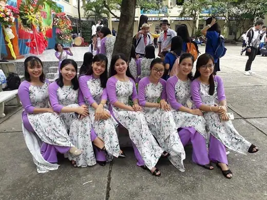 Bảy cô giáo mặc áo dài đồng phục trắng hoa tím ngồi thành hàng trong sân trường.
