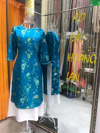 Áo dài cách tân gấm xanh phối chân váy trắng trên ma-nơ-canh, ảnh có chữ tên tiệm.