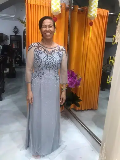 Một phụ nữ lớn tuổi mặc đầm dạ hội ren xám kết cườm đứng trước rèm cam.