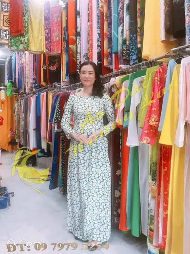 Một phụ nữ mặc áo dài trắng in hoa cúc hoạ mi đứng trong tiệm, ảnh có logo tiệm.