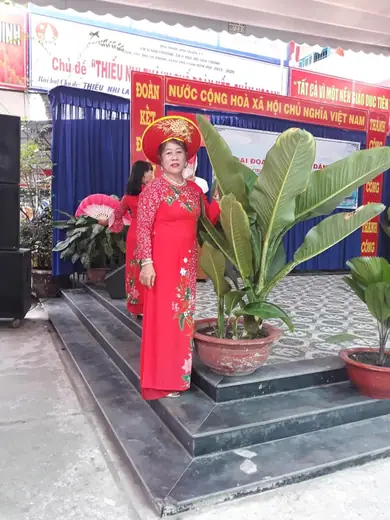 Một phụ nữ mặc áo dài đỏ hồng, đội mấn, đứng trước hội trường treo băng rôn.