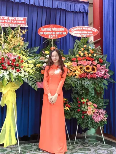 Một cô giáo mặc áo dài cam đứng giữa các lẵng hoa mừng ngày Nhà giáo.