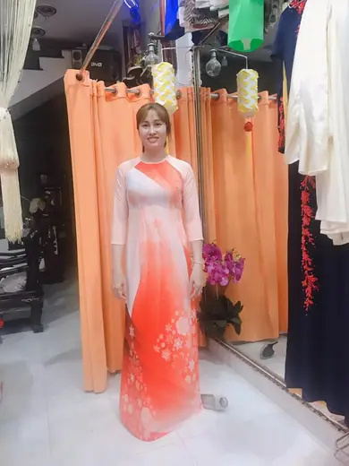 Một phụ nữ mặc áo dài trắng chuyển cam in hoa tuyết đứng trong tiệm.
