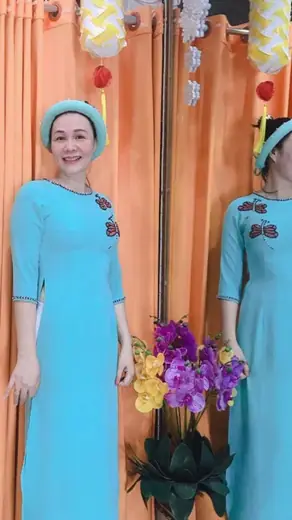 Một phụ nữ mặc áo dài xanh ngọc, đội mấn cùng màu, đứng trước gương.
