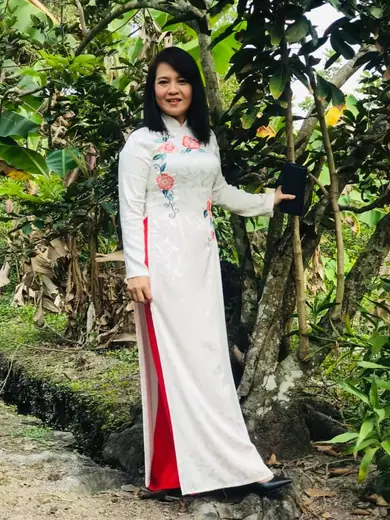 Một phụ nữ mặc áo dài trắng thêu hoa hồng, quần đỏ, đứng trong vườn cây.