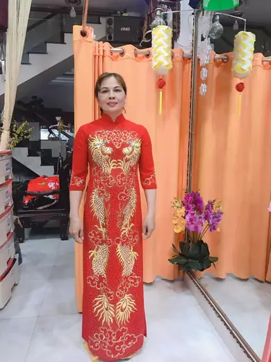 Một phụ nữ mặc áo dài đỏ thêu chim phượng vàng đứng trước rèm cam.