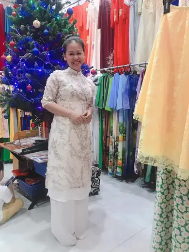 Một phụ nữ mặc áo dài cách tân ren kem đứng trong tiệm cạnh cây thông Noel.