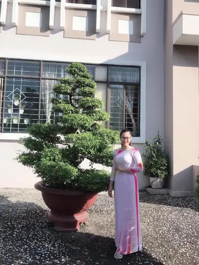 Một phụ nữ mặc áo dài trắng phối hồng đứng cạnh chậu cây cảnh trước toà nhà.