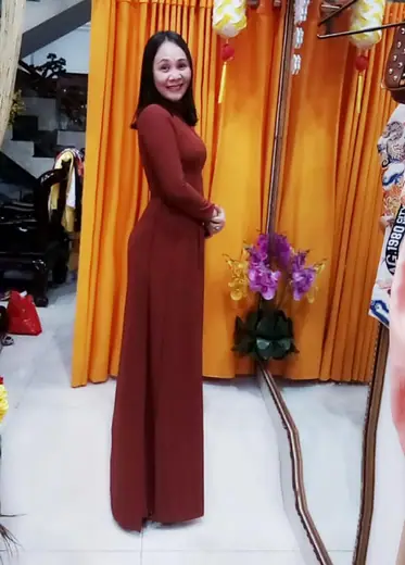 Một phụ nữ mặc áo dài đỏ nâu đứng nghiêng trước rèm cam.