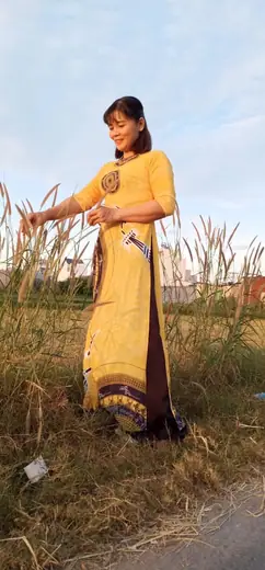 Một phụ nữ mặc áo dài vàng, quần nâu, đứng giữa bãi cỏ lau.