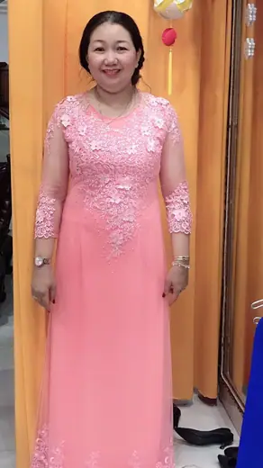Một phụ nữ mặc áo dài hồng đính ren nổi đứng trước rèm vàng.