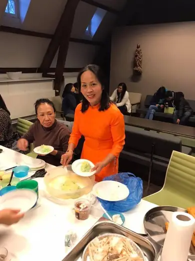 Một phụ nữ mặc áo dài cam đứng bên bàn ăn cùng một cụ bà.
