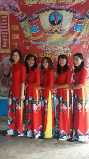 Năm phụ nữ mặc áo dài đồng phục đỏ in hoa đứng trước phông lễ mừng thọ.