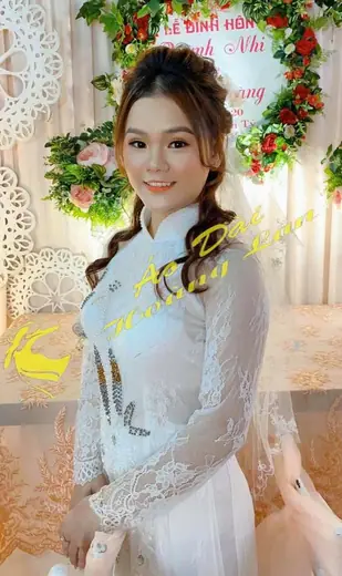 Cô dâu mặc áo dài ren trắng đứng trước phông lễ đính hôn kết hoa, ảnh có logo tiệm.