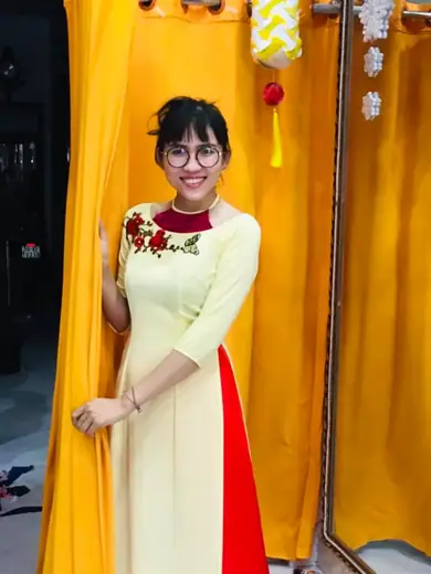 Một cô gái đeo kính mặc áo dài màu kem thêu hoa hồng đỏ, quần đỏ, đứng bên rèm vàng.