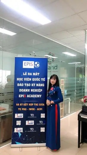 Một phụ nữ mặc áo dài xanh đen đứng cạnh standee lễ ra mắt một học viện đào tạo.