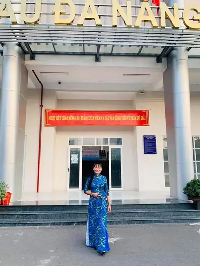 Một phụ nữ mặc áo dài xanh hoạ tiết đứng trước nhà thi đấu.