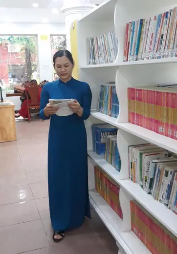 Một cô giáo mặc áo dài xanh đen đứng đọc sách trong thư viện.