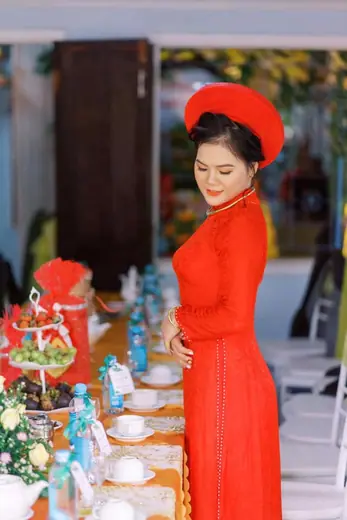 Cô dâu mặc áo dài ren đỏ đứng nghiêng bên bàn tiệc.