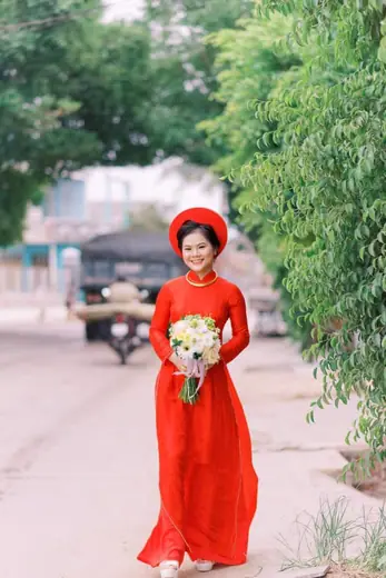 Cô dâu mặc áo dài đỏ ôm bó hoa đứng trên con đường rợp cây.
