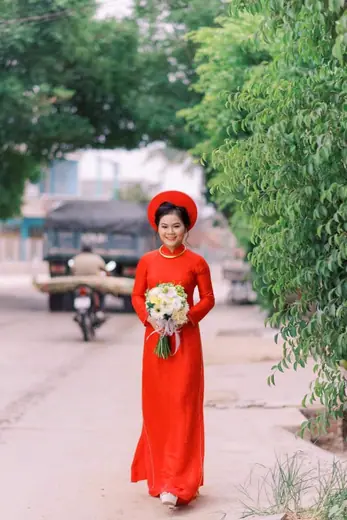Cô dâu mặc áo dài đỏ ôm hoa đứng giữa đường làng.