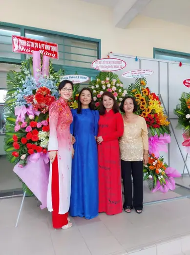 Bốn phụ nữ mặc áo dài trắng đỏ, xanh dương, đỏ và kem đứng giữa các lẵng hoa chúc mừng.