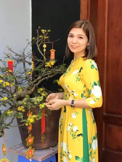 Một phụ nữ mặc áo dài vàng in hoa đứng bên chậu mai Tết.