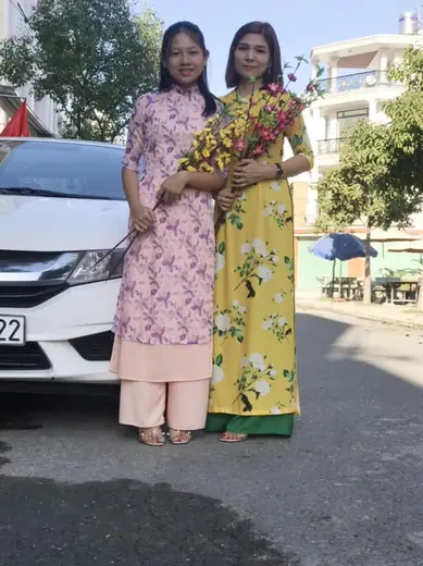 Hai phụ nữ mặc áo dài tím nhạt in hoa và vàng in hoa cầm hoa đứng bên ô tô.