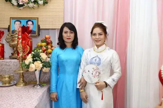 Cô dâu mặc áo dài trắng in hoa xanh đứng cùng một phụ nữ mặc áo dài xanh da trời bên bàn thờ gia tiên.