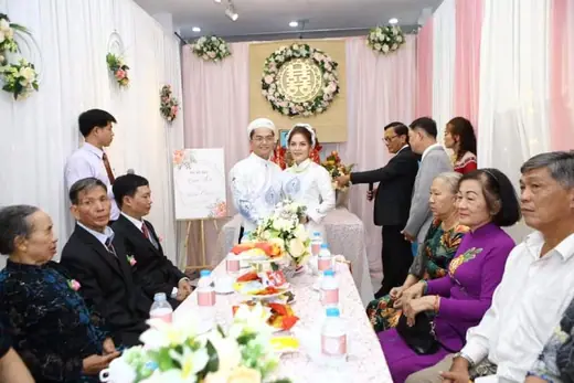 Hai họ ngồi quanh bàn trà trong lễ cưới, cô dâu chú rể mặc áo dài trắng.