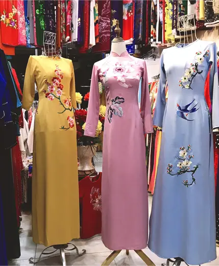 Ba chiếc áo dài vàng mù tạt, hồng đất và xanh nhạt thêu hoa, chim én trên ma-nơ-canh.