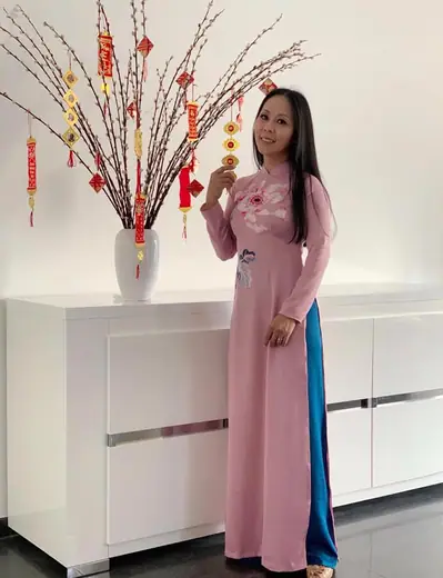 Một phụ nữ mặc áo dài hồng đất, quần xanh, đứng bên bình cành đào trang trí Tết.
