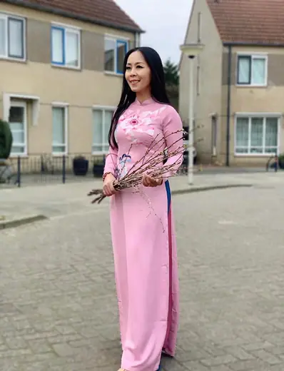 Một phụ nữ mặc áo dài hồng thêu hoa mẫu đơn cầm cành hoa đứng trên phố có những ngôi nhà mái dốc.